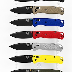 May include: A collection of seven folding knives with black blades and textured handles in black, tan, blue, red, silver, yellow, and olive green. Each knife features a butterfly logo on the blade and a locking mechanism on the handle.