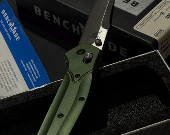 May include: A folding knife with a green handle and a metallic blade featuring the Benchmade logo. The knife is positioned in a black box with the text Benchmade Knife Company. A blue manual with the text Benchmade Use and Care Manual is visible.