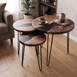 May include: A set of three round nesting tables featuring natural wood grain tops with a black resin inlay and thin black metal hairpin legs. The largest table holds a ceramic cup on a saucer and a book titled Wood & Epoxy.
