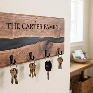 May include: A rectangular wooden key holder featuring a dark brown wood grain and a horizontal black resin inlay. The text THE CARTER FAMILY is engraved in the wood. Four black metal hooks are attached to the bottom, each holding a set of keys.