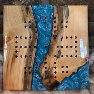 May include: A square wooden board game featuring two natural wood slabs with live edges separated by a central river of swirling blue epoxy resin. The board contains multiple small, dark red circular pegs embedded in a grid pattern across the wood.