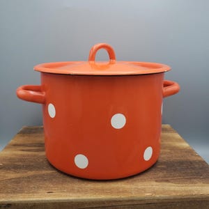 May include: An orange enamel pot with a lid and two side handles. The pot features white polka dots and a matching orange lid with a loop handle. The pot is sitting on a wooden surface.