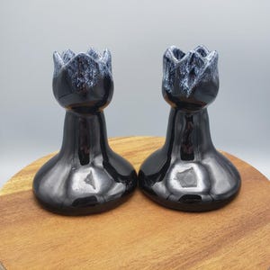 May include: Two black ceramic candle holders with a glossy finish. The top of each holder has a textured, dark blue and white design, resembling a flower. The holders are displayed on a wooden surface.