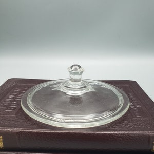 May include: A clear glass lid with a small knob handle. The lid is round and sits atop a stack of two brown books. The glass is transparent, allowing a view of the books below.