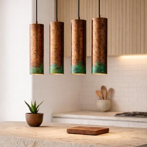 May include: Four cylindrical pendant lights with a copper finish and a green patina at the bottom. The lights hang from black cords in a kitchen setting. A small plant in a wooden pot and a wooden cutting board are on the countertop.