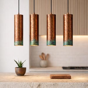 May include: Four copper-finished cylindrical pendant lights with a green patina at the base, suspended from black cords. The lights are in a kitchen setting. A small plant in a wooden pot is on the countertop.