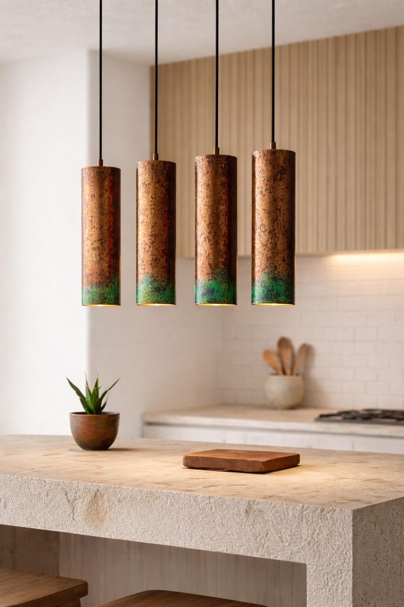 May include: Four cylindrical pendant lights with a copper finish and a green patina at the bottom. The lights hang from black cords in a kitchen setting. A small plant in a wooden pot and a wooden cutting board are on the countertop.