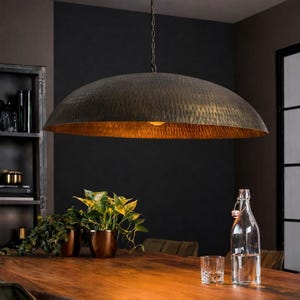 Puede incluir: Una gran lámpara colgante de metal martillado con un interior dorado y cálido cuelga sobre una mesa de madera. La pantalla en forma de cúpula está suspendida por una cadena. La habitación presenta una pared oscura y una estantería.