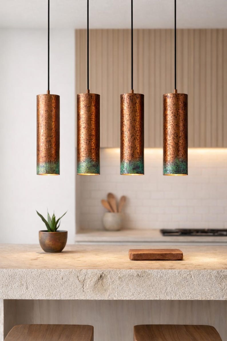 May include: Four copper-finished cylindrical pendant lights with a green patina at the base, suspended from black cords. The lights are in a kitchen setting. A small plant in a wooden pot is on the countertop.