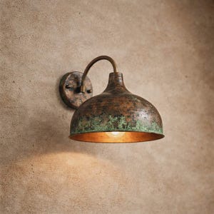 Może przedstawiać: Rustykalna lampa ścienna zewnętrzna. Oprawa posiada miedziany, kopulasty klosz z zieloną patyną, zakrzywione ramię i okrągłą płytę montażową. Światło jest włączone, emitując ciepłą poświatę.