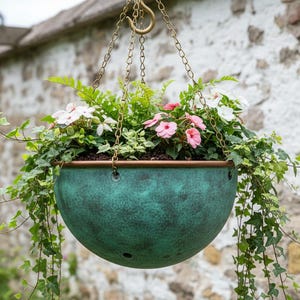 May include: A hanging planter with a verdigris finish, filled with green foliage and pink and white flowers. The planter is suspended by brass-coloured chains against a stone wall.