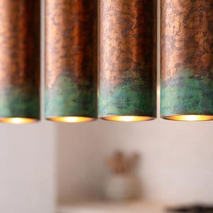 May include: Four copper pendant lights with a green patina at the base, hanging from the ceiling. The lights are cylindrical and illuminated from within, creating a warm glow. The background is blurred, suggesting a kitchen or dining area.