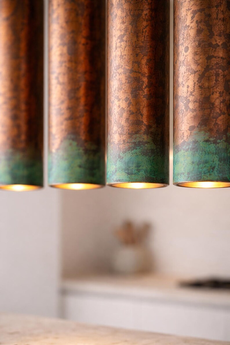 May include: Four copper pendant lights with a green patina at the base, hanging from the ceiling. The lights are cylindrical and illuminated from within, creating a warm glow. The background is blurred, suggesting a kitchen or dining area.