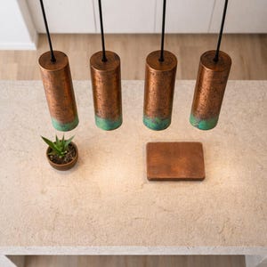 May include: An aerial view of a kitchen island featuring four copper pendant lights with a green patina, suspended from black cords. A small plant and a wooden tray sit on the island. The lights are cylindrical.