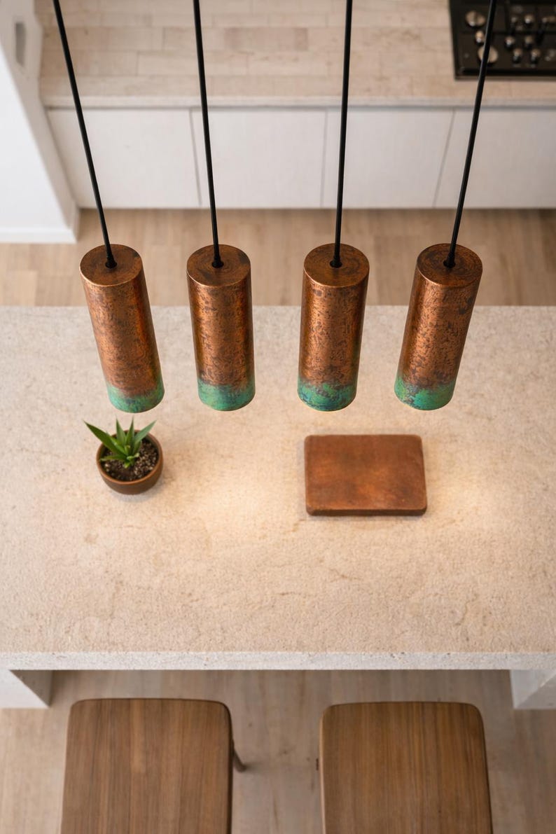 May include: An aerial view of a kitchen island featuring four copper pendant lights with a green patina, suspended from black cords. A small plant and a wooden tray sit on the island. The lights are cylindrical.