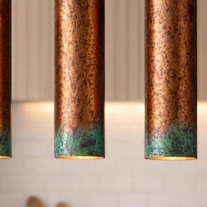 May include: Three copper-finished pendant lights with a green patina at the base. The cylindrical lights hang from the ceiling, providing illumination. The copper has a textured finish. The lights are suspended above a kitchen area.