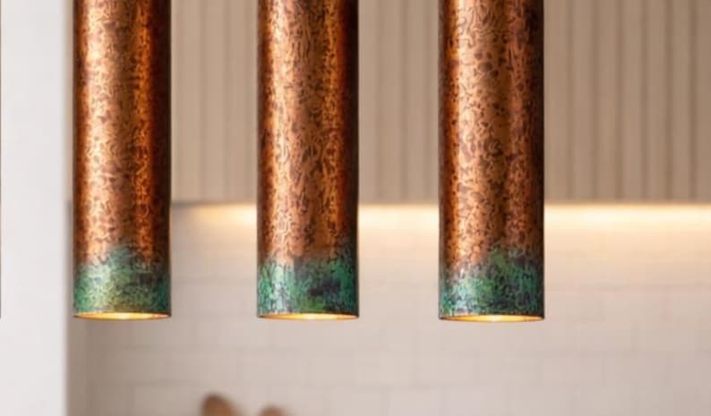 May include: Three copper-finished pendant lights with a green patina at the base. The cylindrical lights hang from the ceiling, providing illumination. The copper has a textured finish. The lights are suspended above a kitchen area.