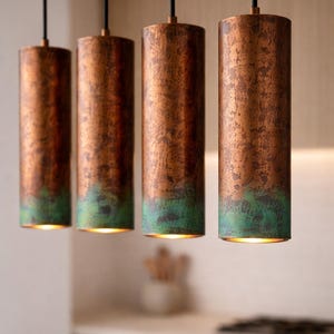 May include: Four copper-finished cylindrical pendant lights with a green patina at the base, suspended from black cords. The lights emit a warm glow from within. These lights are a decorative feature, suitable for a kitchen or dining space.