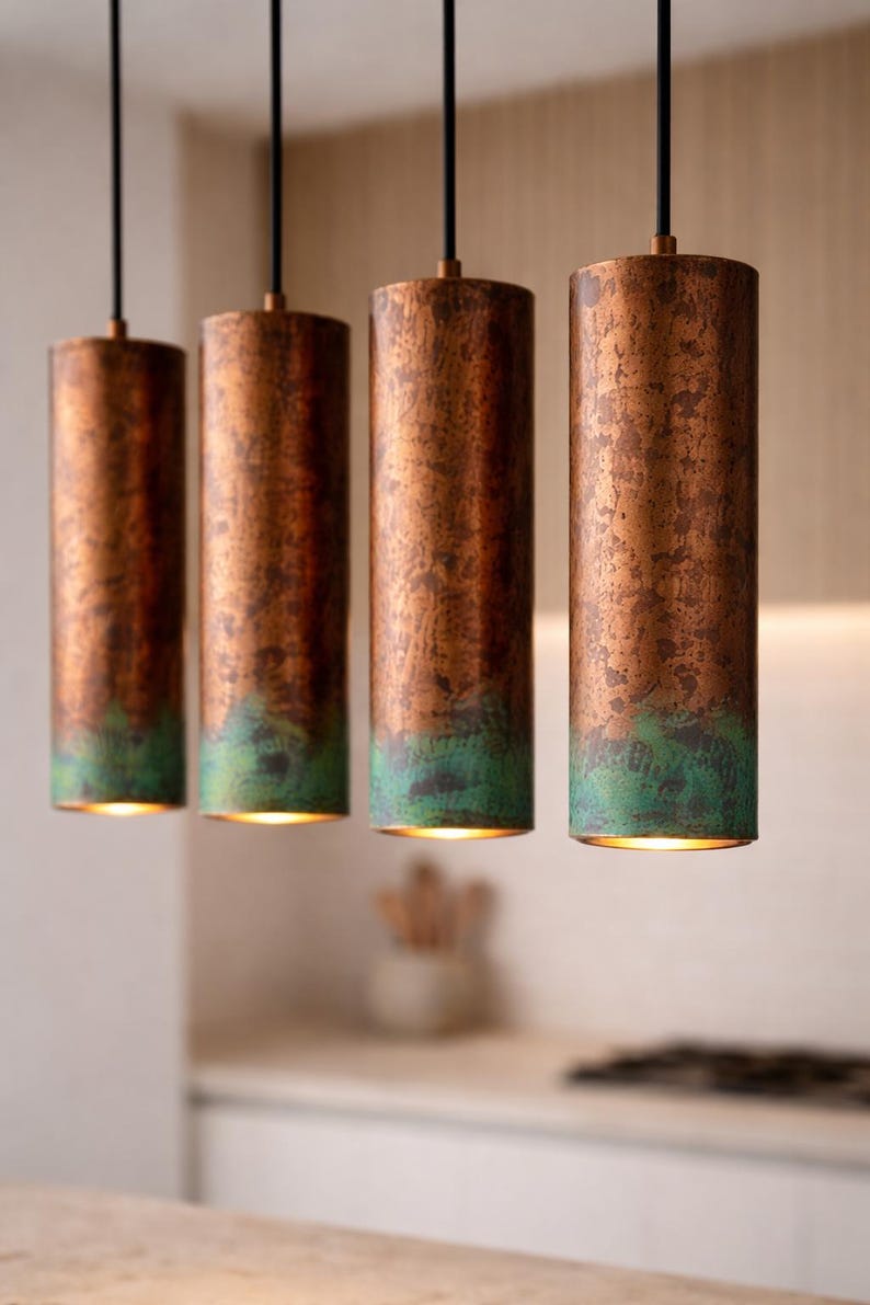 May include: Four copper-finished cylindrical pendant lights with a green patina at the base, suspended from black cords. The lights emit a warm glow from within. These lights are a decorative feature, suitable for a kitchen or dining space.
