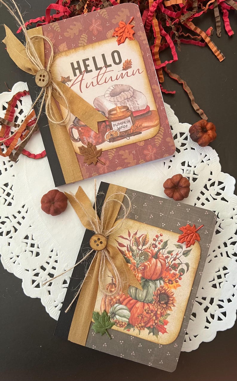 May include: Two small notebooks with fall-themed covers. The top notebook has a red cover with a brown and white sweater, a candle, and the text "Hello Autumn". The bottom notebook has a gray cover with a stack of pumpkins, sunflowers, and fall leaves.