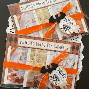 May include: Two "Witches Brew Tea Sampler" packs. Each pack features tea bags with flavors like "Salted Caramel" and "Hot Cinnamon." They are decorated with orange ribbons, a bat, and a "Black Cat Tea Co." tag, perfect for Halloween.