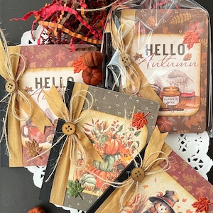 May include: Three small journals with fall-themed covers. The covers feature pumpkins, leaves, and a scarecrow. The journals are tied with twine and decorated with fabric bows. The journals are on a white lace doily.