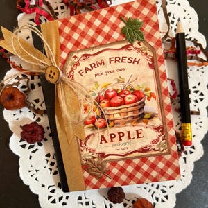 May include: A decorative notebook with a red and white checkered pattern and a label that reads "Farm Fresh Apple Orchard". The label features a basket of red apples. The notebook is tied with twine and a wooden button.
