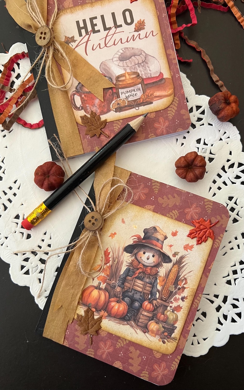 May include: Two small notebooks with fall-themed covers. The top notebook has a brown cover with the text "HELLO Autumn" and a fall-themed illustration. The bottom notebook has a red cover with a scarecrow illustration and the text "Pumpkin Spice".