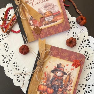 May include: Two small notebooks with fall-themed covers. The top notebook has a brown cover with the text "HELLO Autumn" and a fall-themed illustration. The bottom notebook has a brown cover with a scarecrow illustration and pumpkins.