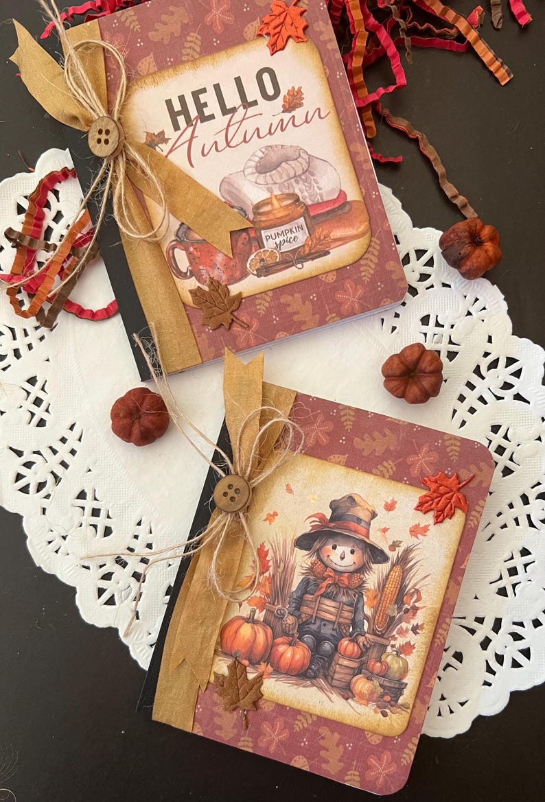 May include: Two small notebooks with fall-themed covers. The top notebook has a brown cover with the text "HELLO Autumn" and a fall-themed illustration. The bottom notebook has a brown cover with a scarecrow illustration and pumpkins.