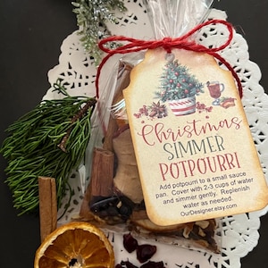 May include: A clear cellophane bag tied with red twine, containing a Christmas simmer potpourri. The tag reads "Christmas Simmer Potpourri" with instructions. Includes dried orange slices, cinnamon sticks, and cranberries. The background features a white doily and evergreen sprigs.