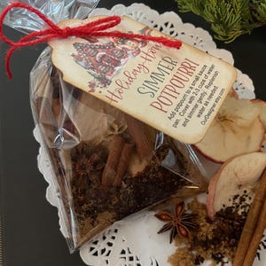 May include: A clear cellophane bag tied with red twine, containing a simmer potpourri mix. The mix includes cinnamon sticks, star anise, and dried apple slices. A tag reads "Holiday How SIMMER POTPOURRI".