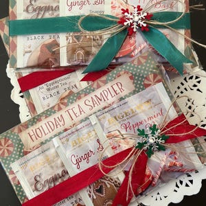 May include: Holiday tea sampler gift sets. Each set includes tea bags of various flavors, such as Eggnog, Ginger Snappy, and Peppermint Bark. The sets are decorated with red and green ribbons, twine, and snowflake ornaments. The packaging has a festive holiday theme.