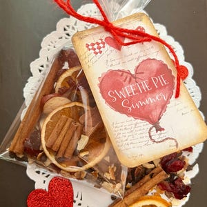 May include: A clear cellophane bag filled with dried orange slices, cinnamon sticks, and other potpourri ingredients. A tag with a red heart and the words "Sweetie Pie Simmer" is attached with red twine. A red glitter heart is also present.