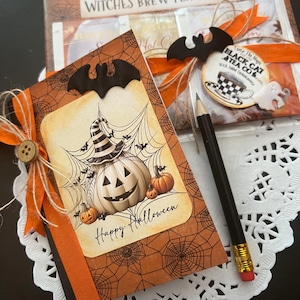 May include: A Halloween-themed gift set featuring a decorative card with a pumpkin and the words "Happy Halloween." The set includes tea samplers, a pencil, and bat-shaped embellishments, all tied with orange ribbon.