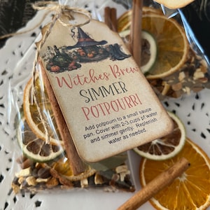 May include: A clear cellophane bag tied with twine, containing dried orange slices, cinnamon sticks, and other potpourri ingredients. A tag reads "Witches Brew Simmer Potpourri" with instructions and a witch-themed illustration.