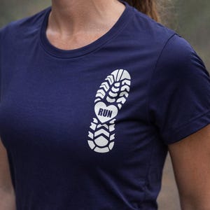 Könnte beinhalten: Marineblaues T-Shirt mit weißem Fußabdruck-Design. Der Fußabdruck enthält das Wort "RUN" in Herzform. Das Shirt ist aus einem weichen Material.