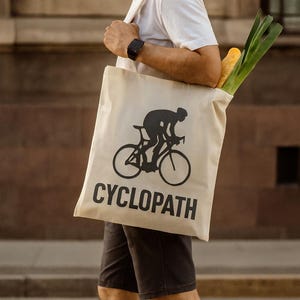 Puede incluir: Bolsa de mano beige con un gr&aacute;fico de ciclista negro y la palabra "CYCLOPATH" en letras negras. La bolsa contiene comestibles, incluyendo una baguette y verduras verdes. La persona lleva una camiseta blanca y pantalones cortos negros.