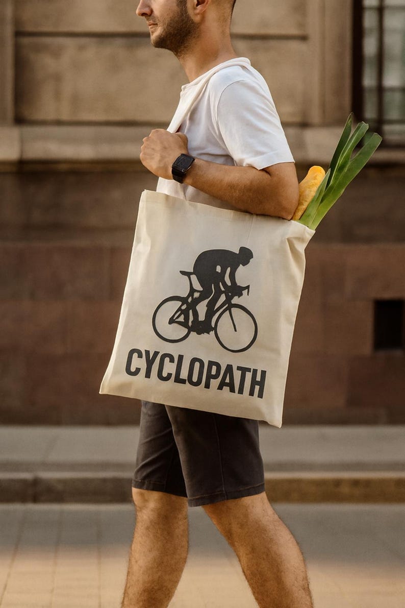 Puede incluir: Bolsa de mano beige con un gr&aacute;fico de ciclista negro y la palabra "CYCLOPATH" en letras negras. La bolsa contiene comestibles, incluyendo una baguette y verduras verdes. La persona lleva una camiseta blanca y pantalones cortos negros.