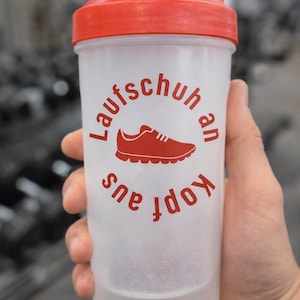 May include: A clear plastic shaker bottle with a red lid. The bottle has a red graphic of a running shoe and the words "Laufschuh an Kopf aus" in red. The bottle is held by a person in a gym setting.