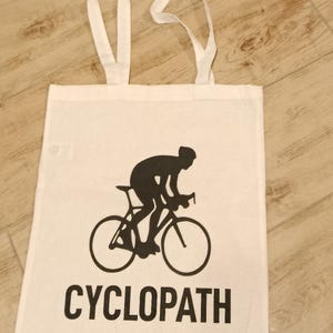 Puede incluir: Bolso de mano blanco con una silueta negra de un ciclista en bicicleta. La palabra "CYCLOPATH" est&aacute; impresa en letras negras en negrita debajo de la imagen. El bolso tiene dos asas blancas.