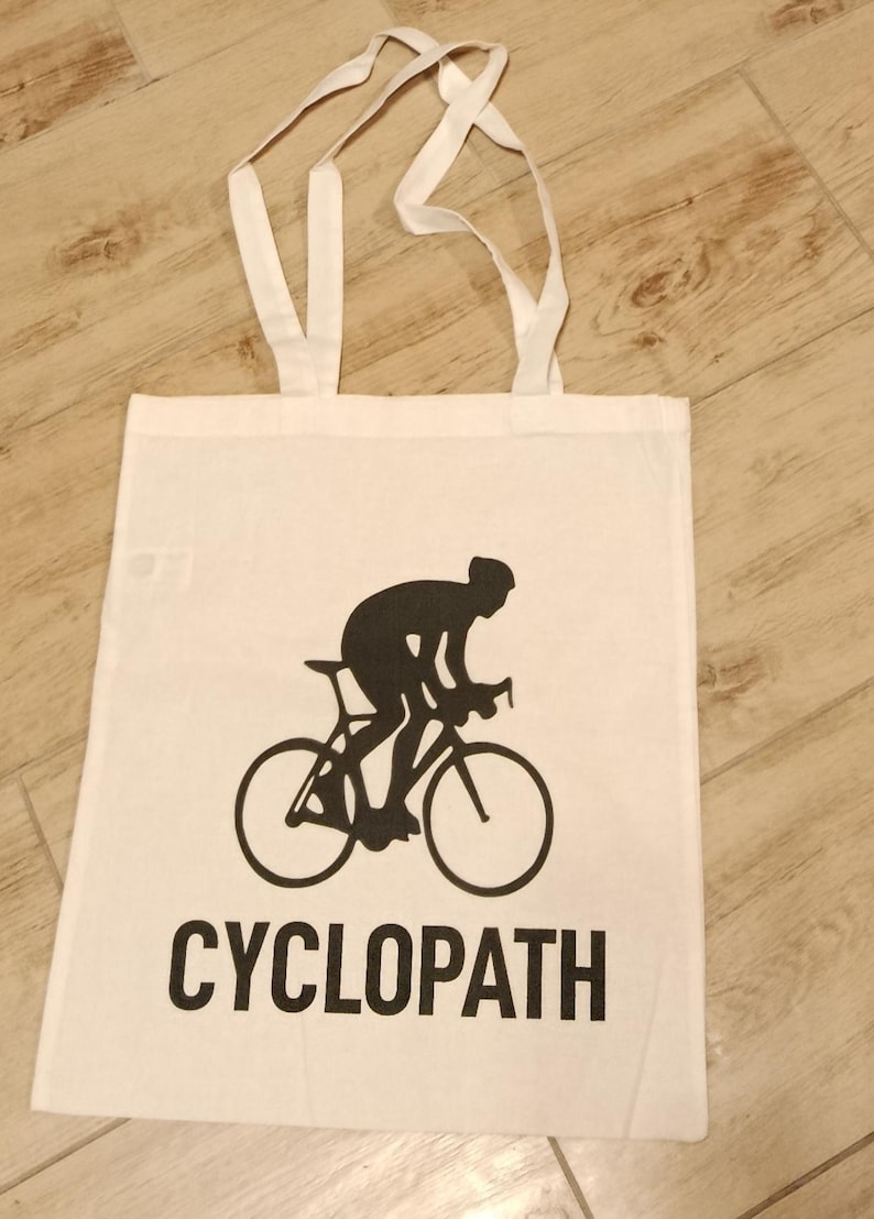 Puede incluir: Bolso de mano blanco con una silueta negra de un ciclista en bicicleta. La palabra "CYCLOPATH" est&aacute; impresa en letras negras en negrita debajo de la imagen. El bolso tiene dos asas blancas.