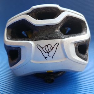 Könnte beinhalten: Weiß-schwarzer Fahrradhelm mit schwarzem Innenraum. Der Helm hat eine schwarz-weiße Handgesten-Grafik auf der Rückseite. Der Helm hat mehrere Belüftungsöffnungen und eine Dreheinstellung.