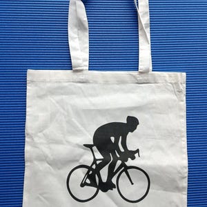 Puede incluir: Bolso tote blanco con una silueta negra de un ciclista en bicicleta. La palabra "CYCLOPATH" est&aacute; impresa en negro debajo. El bolso tiene dos asas blancas y est&aacute; sobre un fondo azul.
