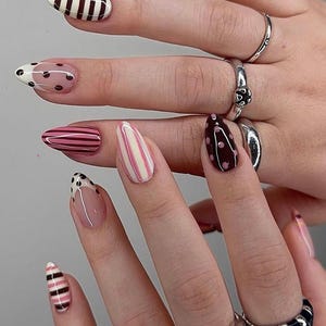 May include: A hand displaying long, almond-shaped nails with diverse designs. The nails feature stripes, polka dots, and French tips, in pink, white, brown, and yellow. The fingers are adorned with silver rings, showcasing a variety of styles and textures.