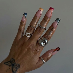 May include: A hand displaying long, square-shaped nails, each with a unique colour gradient. The nails have a pink base with colourful tips in shades of orange, red, and green. Several silver rings are worn on the fingers, and a butterfly tattoo is visible on the wrist.