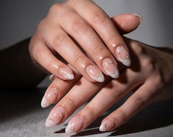 May include: Oval-shaped nails with a clear base, white tips, and gold star and dot accents. The nails are adorned with small gold stars and dots, creating a delicate and elegant design. The manicure is set against a neutral background.