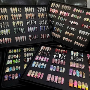 Peut inclure: Trois plateaux de présentation noirs remplis d'une vaste collection de faux ongles à coller de formes, longueurs et motifs variés, incluant des couleurs unies, des paillettes, des motifs floraux, des strass et des dessins graphiques peints à la main.
