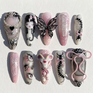 Puede incluir: Un juego de diez uñas postizas con diseños variados que incluyen un corazón con perlas, un rostro estilizado con lazo, una mariposa negra, arte lineal, un ciempiés, una cabra de tres ojos, múltiples ojos y un marco de cinta rosa.