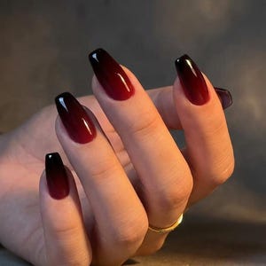 Puede incluir: Una mano con uñas largas en forma de ataúd. Las uñas presentan un diseño degradado, pasando de un borgoña oscuro en la base a un negro brillante en las puntas. Un anillo dorado adorna el dedo anular. El fondo es gris neutro.